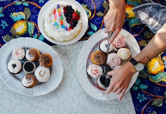 ruce krájí na větším talíři muffin na půl, vedle jsou další talíře s dobrotami